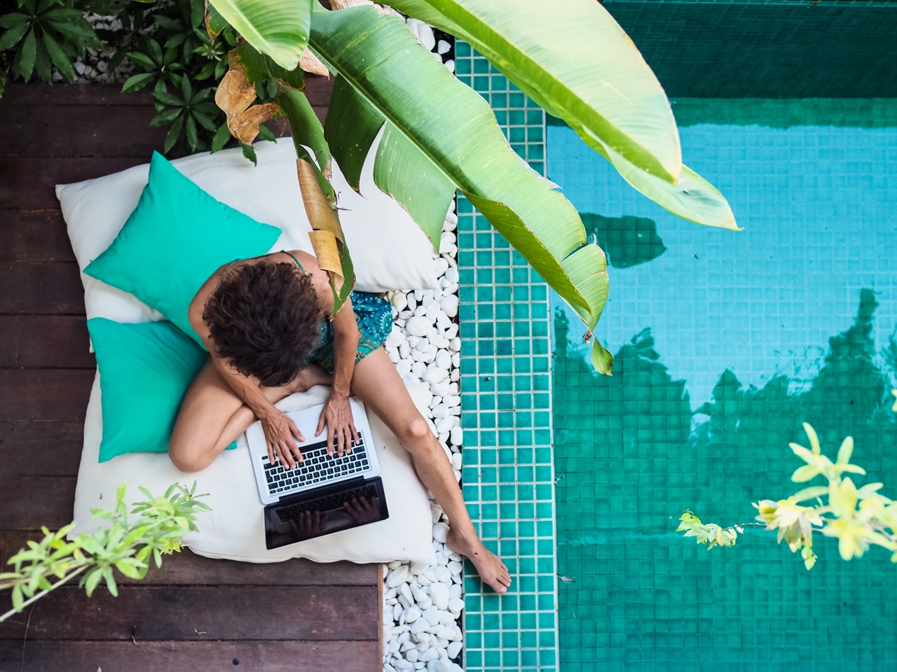 Aerial view of person working beside a pool wearing blue swim shorts and sitting on white and teal colored pillows
